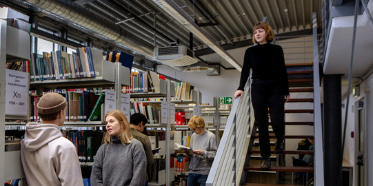 Two students stand talking next to bookshelves. One student walks down the stairs.
